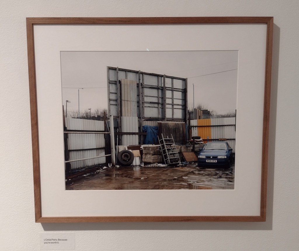 A photograph showing the back of a billboard and a junk yard with an old car, some shelving units and a tyre